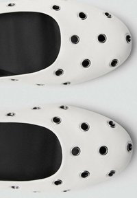Weiße Schuhe aus weichem Material mit großen runden Belüftungsöffnungen und metallischen Akzenten. Das schwarze Innenfutter ergänzt das Design.