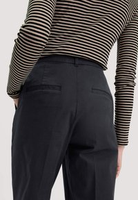 Zwarte op maat gemaakte broeken met een soepele textuur, voorzien van twee achterzakken. De bovenkant is een lang mouw, beige en zwart gestreept shirt.