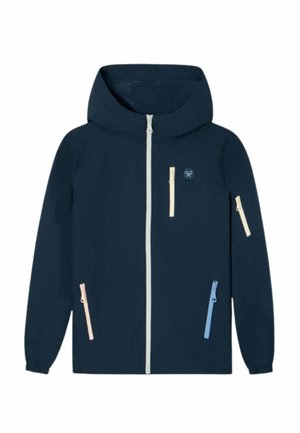 Niño caminando con una chaqueta azul marino, jersey crema sobre una camisa azul, vaqueros azul claro y zapatillas oscuras contra un fondo liso.