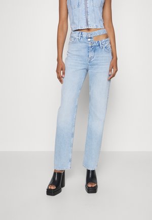 Vrouw draagt lichtblauwe spijkerbroek met uitsparing en dubbele tailleband, gecombineerd met zwarte open teens plateauhakken en een mouwloos denim cropped top.