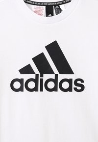 Camiseta blanca de Adidas con un gran logo negro de Adidas y texto centrado en el pecho, etiquetas del collar visibles en el interior del escote.