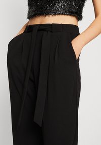 Pantalon noir ajusté taille haute, avec ceinture nouée, poches latérales et détails plissés pour une texture supplémentaire.