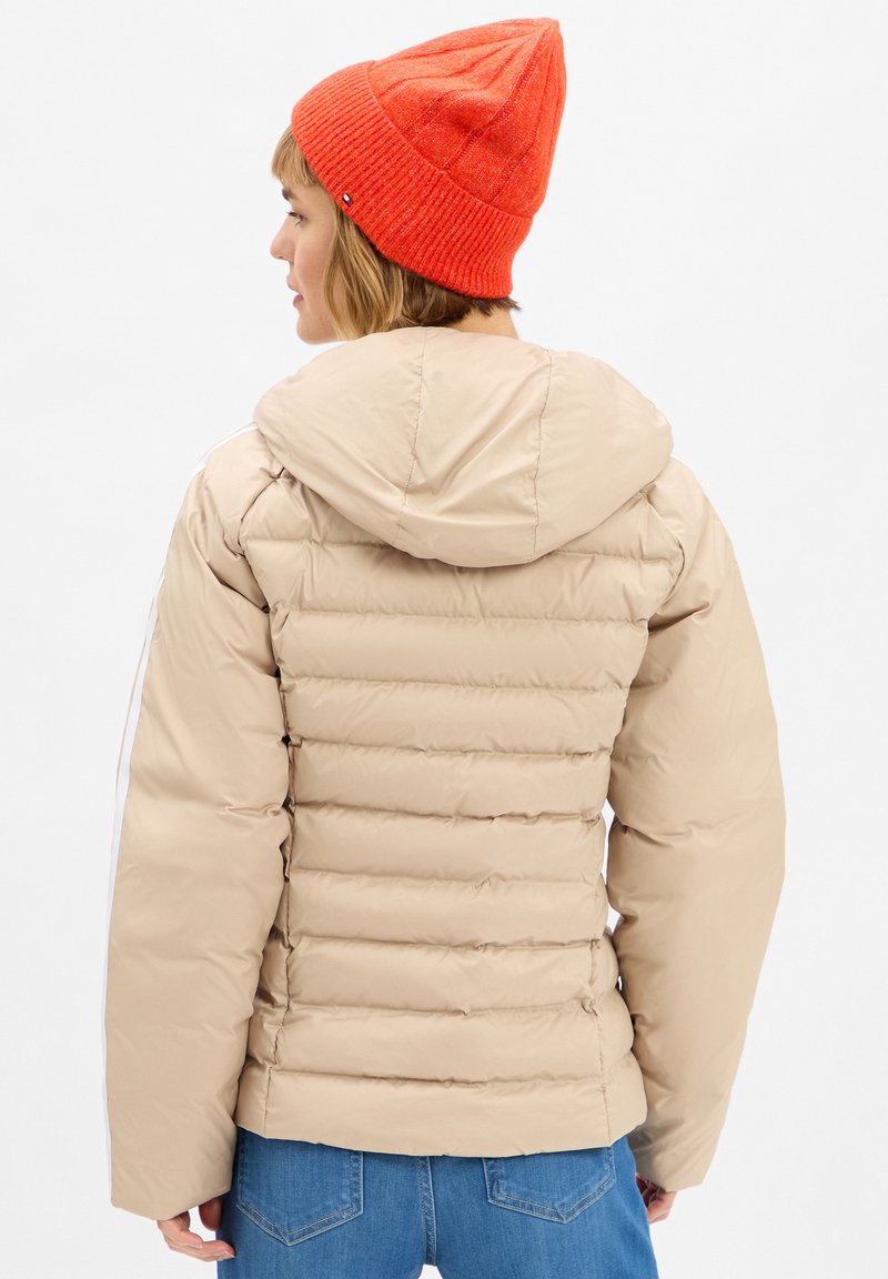 Persona che indossa un piumino beige con cappuccio e un berretto di lana arancione, in piedi con la schiena voltata, jeans blu visibili.