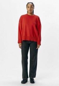 Rode oversized trui met geribbelde manchetten, gecombineerd met hooggeguard donkerdenim en chunky zwarte laarzen. Gladde textuur en relaxte pasvorm.