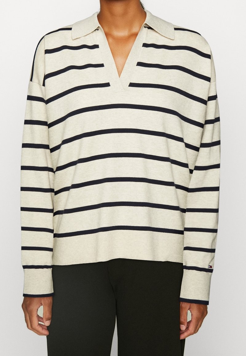 Pull crème à manches longues avec des rayures horizontales noires et un col en V, porté avec un pantalon noir.