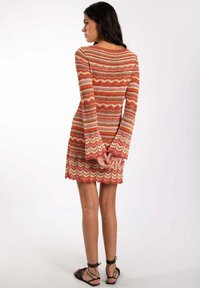 Robe tricotée avec de longues manches évasées dans des teintes chaudes d'orange, de rose et de crème, présentant un motif en zigzag ondulé ; longueur genou avec un ourlet à volants.