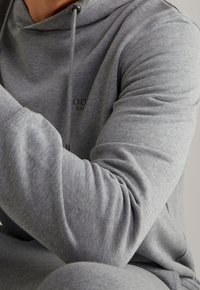 Sudadera gris con cuello de cordón, puños acanalados y textura suave. Presenta un pequeño logo en la zona del pecho. Diseño de corte casual.