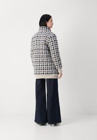 Oversized Wollmischungsmantel in Schwarz, Weiß und Beige mit Hahnentrittmuster. Verfügt über einen hohen Kragen und gerippte Bündchen, kombiniert mit weit geschnittenen Jeanshosen.