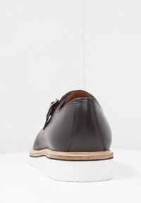 Scarpa in pelle nera con finitura liscia, punta arrotondata e suola color marrone chiaro; presenta una suola bianca, leggermente rialzata e un dettaglio con fibbia.