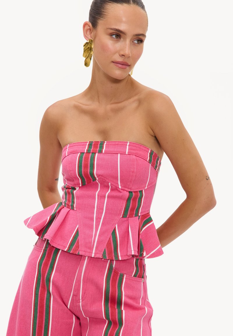 Femme portant un top peplum rose sans bretelles et un pantalon assorti avec des rayures verticales blanches, vertes et rouges, accompagné de grandes boucles d'oreilles dorées.