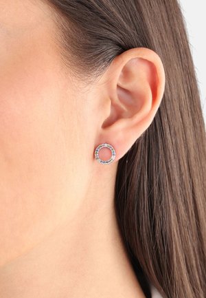 Gros plan sur l'oreille d'une femme portant une petite boucle d'oreille clou circulaire avec des pierres précieuses multicolores, et des cheveux bruns raides repliés derrière l'oreille.