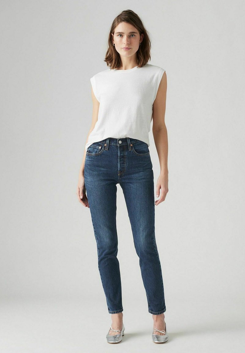 Chemise sans manches blanche associée à un jean taille haute bleu foncé et des ballerines argentées, présentant un design simple et une coupe classique.