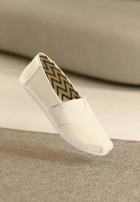Valkoinen kangaskengä, joka on slip-on-tyyppinen ja jossa on pyöristetty varvas. Pohjan tekstuuri on kermaisen värinen. Sisäpuolella on ruskea- ja mustakuvioinen zigmalle.