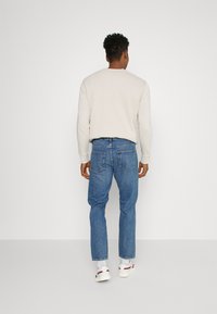 Jasno beżowa bluzka z długim rękawem połączona z niebieskimi dżinsami o prostym kroju i białymi trampkami. Dżinsy mają tylne kieszenie i są lekko luźne.