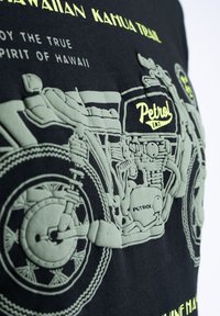 Černé tričko s embosovaným designem motocyklu v zelené barvě a textem "Petrol" spolu s havajskou frází, zdůrazňující složité detaily a texturu.