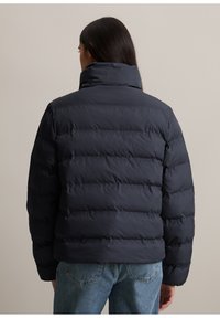 Doudoune marine avec col montant et design matelassé. Tissu doux et texturé avec des poignets élastiques et une silhouette ajustée.