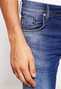 Blå denimjeans med en klassisk design som har subtila blekningar, femficksstil och kontrasterande orange sömmar. Hand som vilar på tyget.