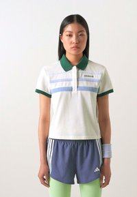 Camisa polo branca com colarinho verde, apresentando riscas horizontais azuis. Combinada com calções azul-marinho, leggings verdes claras e uma bracelete.