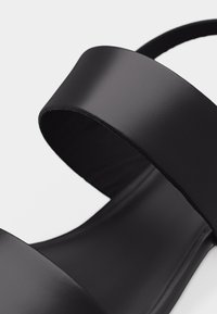 Gros plan sur la bride de sandale noire en matériau lisse semblable au cuir, avec une large bande épaisse sur le dessus du pied.