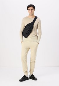Bolso de nylon negro tipo bandolera con bolsillos con cremallera, llevado en diagonal. Modelo con una camisa de manga larga beige y pantalones de chándal a juego, zapatillas negras.