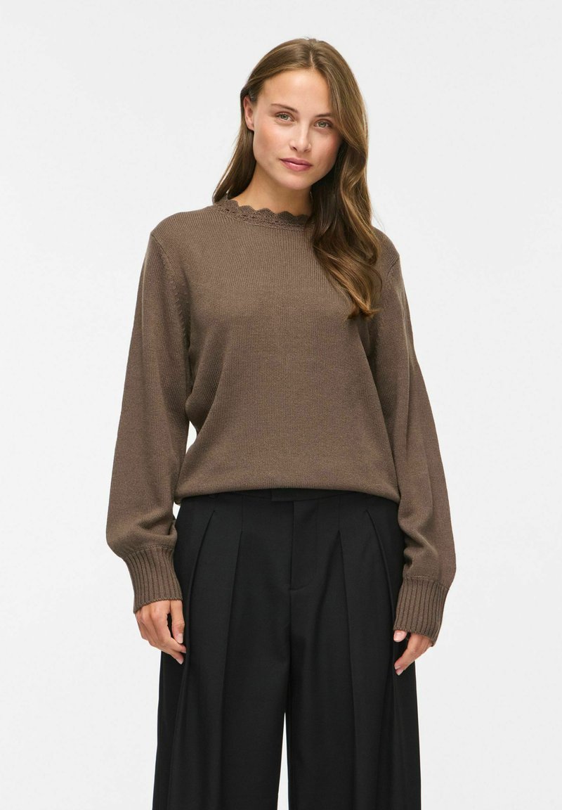 Femme aux longs cheveux bruns portant un pull en maille marron à manches longues et un pantalon noir plissé taille haute, debout devant un fond blanc.