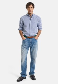 Camisa de botón a rayas azul claro, jeans azules, cinturón de cuero marrón y zapatos negros, con una bufanda estampada alrededor del cuello.