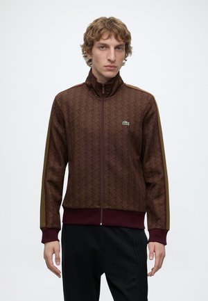 Junger Mann mit lockigem Haar trägt eine braune gemusterte Reißverschlussjacke mit olivgrünen Streifen an den Ärmeln und dunkle Hosen vor einem einfarbigen Hintergrund.