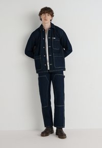 Denim jacket and pants in dark blue, featuring contrasting stitching, pockets, and a relaxed fit. Brown boots complete the look.