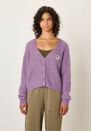 Cardigan lilas en matériau moelleux, avec un décolleté en V profond, six boutons et une patch en forme de cœur. Associé à un pantalon vert olive.