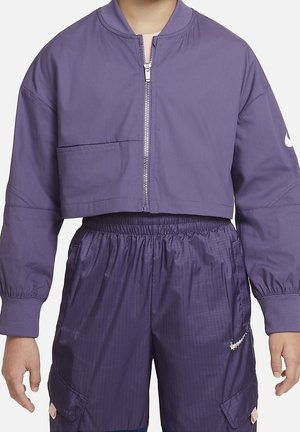 Person iført en kort lilla lynlåstrøje og matchende lilla sportsskjorter med elastisk talje og Nike-logo på ærmet og buksebenet.