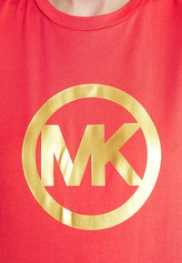 Červené bavlněné tričko s velkým lesklým zlatým logem "MK" uzavřeným v kruhovém zlatém orámování. Hladká textura a kulatý výstřih.