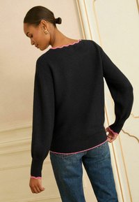 Čierny sveter s textúrovaným povrchom, zdobený vlnitým ružovým lemom na výstrihu a rukávoch. Skombinovaný s modrými džínsami.