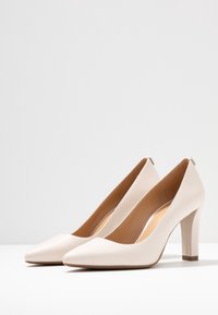Des chaussures à talons hauts en cuir verni beige avec un bout pointu et un talon carré de 8,9 cm. Texture lisse, design minimaliste et sans embellissements visibles.