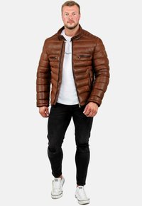 Bruine leren pufferjack met een gequiltde textuur, ritszakken en hoge kraag, gecombineerd met een wit T-shirt en zwarte versleten jeans.