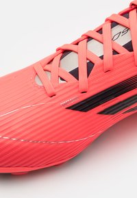 Botas de fútbol de un vibrante color coral con textura acanalada y acentos en negro. Presentan un diseño de cordones, la marca "Fiberskin" y una forma aerodinámica.