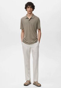 Olivově zelená polo košile s krátkým rukávem, zkombinovaná s světlými béžovými lněnými kalhotami a tmavými sandály, vystavená na neutrálním pozadí.