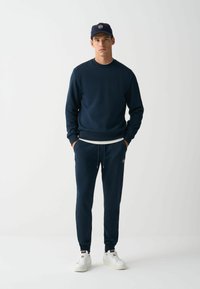 Temno modra trenirka in hlače, z ribastimi manšetami, pasom na vrvico in belimi detajli, v kombinaciji z belimi supergami in temno modrim kapucarjem.