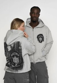 Grau Kapuzenpullover mit Reißverschluss, die grafische Drucke auf der Vorder- und Rückseite haben. Das Material ist weich, mit einer lockeren Passform und Kängurutaschen.