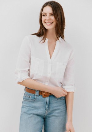Chemise blanche en lin à manches retroussées, dotée de deux poches avant. Associée à un jean en denim bleu clair et une ceinture marron.