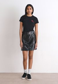 T-shirt in cotone nero con ricami rossi, abbinato a una mini gonna in pelle nera con doppia fibbia sul davanti, indossata con sneakers nere e calzini bianchi.