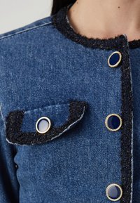 Jachetă din denim cu un tăietor texturat negru, având un buzunar cu un buton decorativ albastru și accente aurii pe butoane.