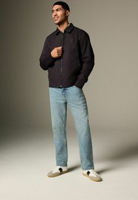 Schwarze Zip-Jacke mit Kragen, kombiniert mit hellblauen Jeans und weißen Sneakers mit grauen und schwarzen Akzenten. Glattes Material und lockere Passform.