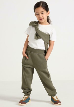 Kinderoutfit, das ein weißes T-Shirt, olivgrüne Jogginghose mit Taschen und buntes Sneakers umfasst. Pullover um die Taille gebunden.