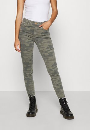 Vrouw draagt groene camouflerende skinny jeans, zwarte veterlaarzen en een wit mouwloos topje, staand met één hand in de zak.