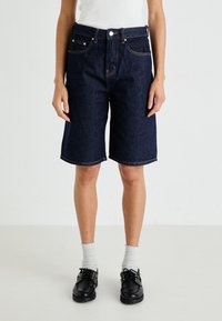 Denim bermudashorts med mørk vask, fem lommer, beltehemper og avslappet passform, kombinert med svarte snørestøvler og lyse sokker.