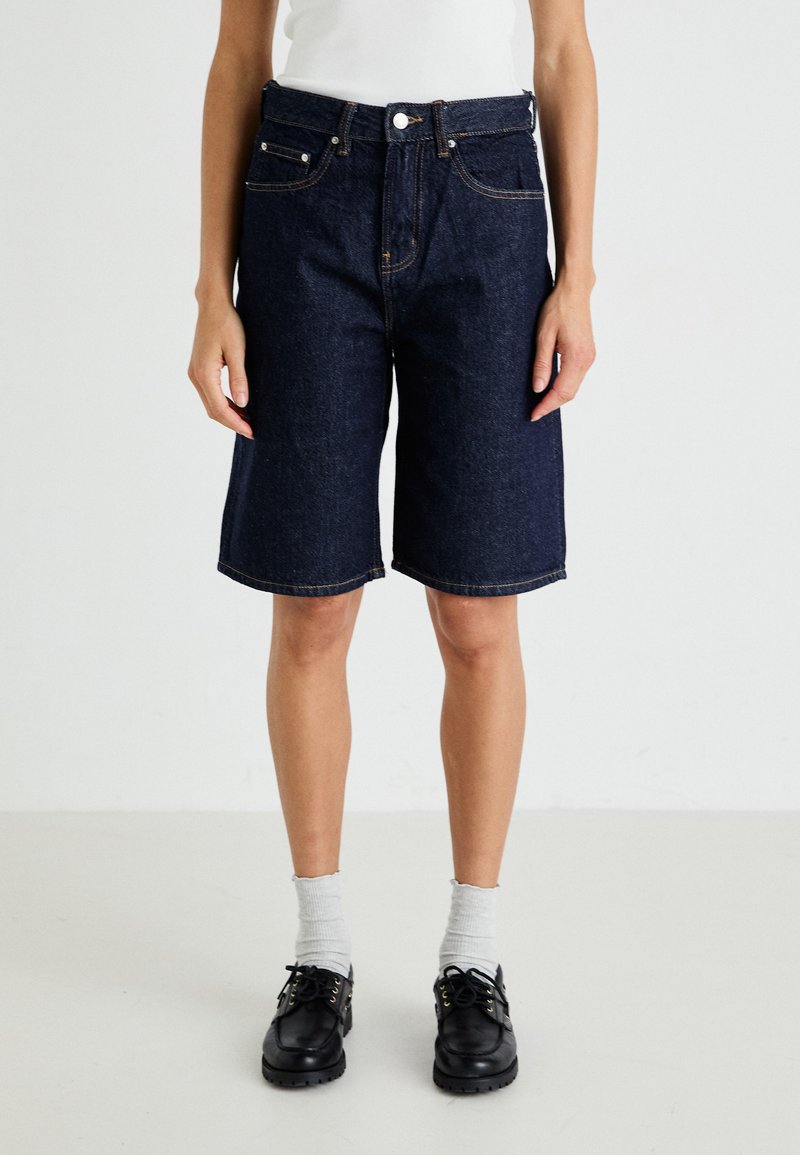 Denim bermudashorts med mørk vask, fem lommer, beltehemper og avslappet passform, kombinert med svarte snørestøvler og lyse sokker.