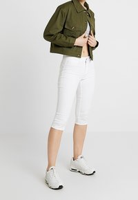 Veste cropped vert olive avec deux poches avant, pantalon capri blanc et baskets blanches. Les caractéristiques incluent une fermeture à boutons et un design ajusté.