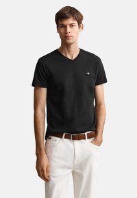 Mladý muž nosí priliehavé čierne tričko s výstrihom do V s malým logom a biele nohavice s hnedým opaskom, stojí s jednou rukou v vrecku.