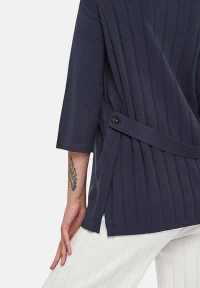 Top a coste di colore blu navy con maniche a lunghezza del gomito, dettagli di bottoni laterali, spacco sull'orlo, indossato sopra pantaloni bianchi strutturati.