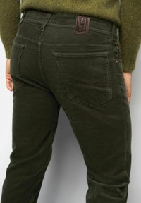 Pantalons en velours côtelé vert olive avec une surface texturée, arborant une étiquette en cuir avec "M5" et deux poches arrière avec des accents brodés.